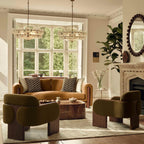 Two white glass disc chandeliers hang side by side in front of a large bay window, over a camel sofa and two olive boucle chairs; Clérachic.