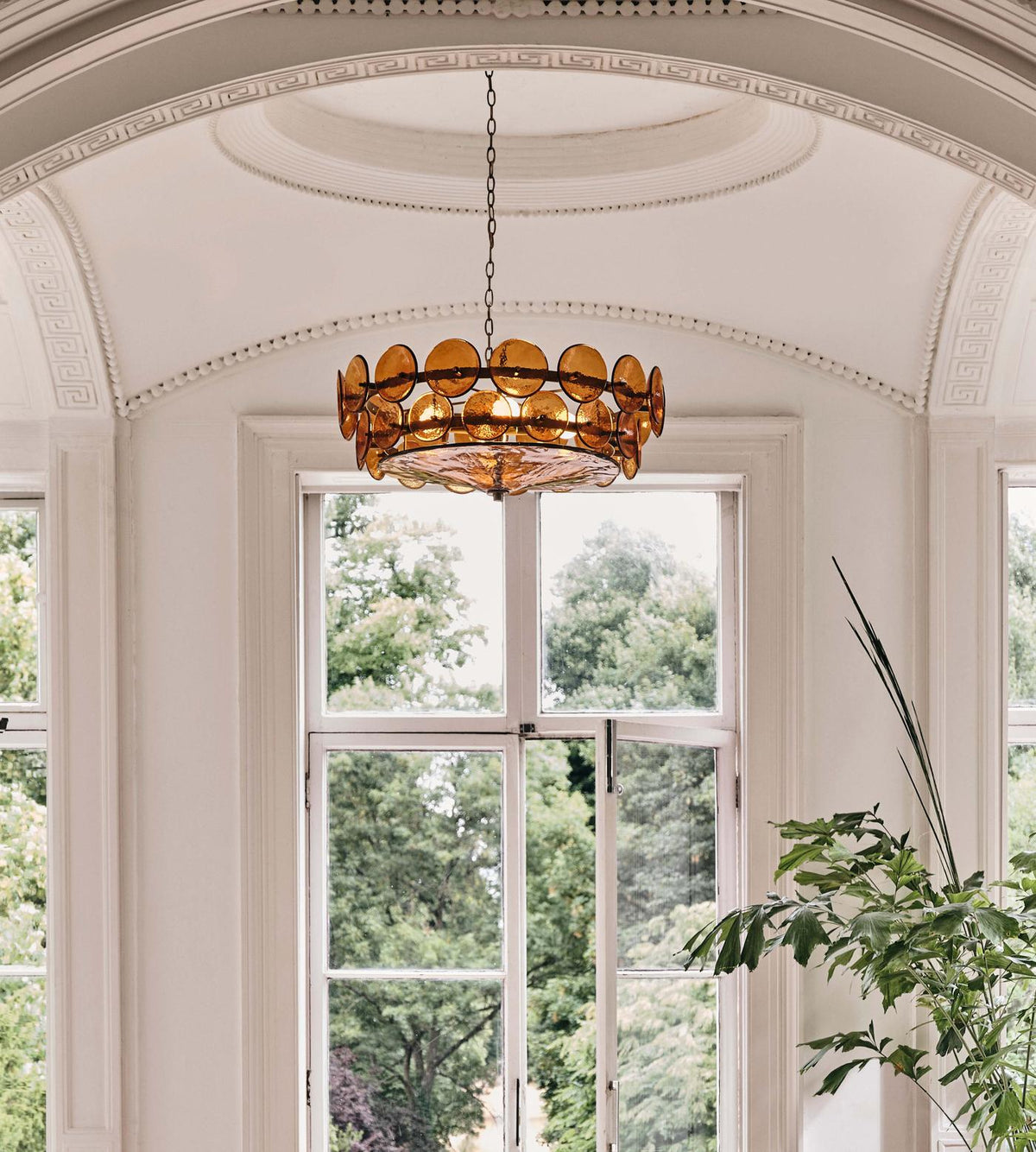 Amber glass disc chandelier from Clérachic framed by a tall arched window and a domed plaster ceiling, with treetops outside.
