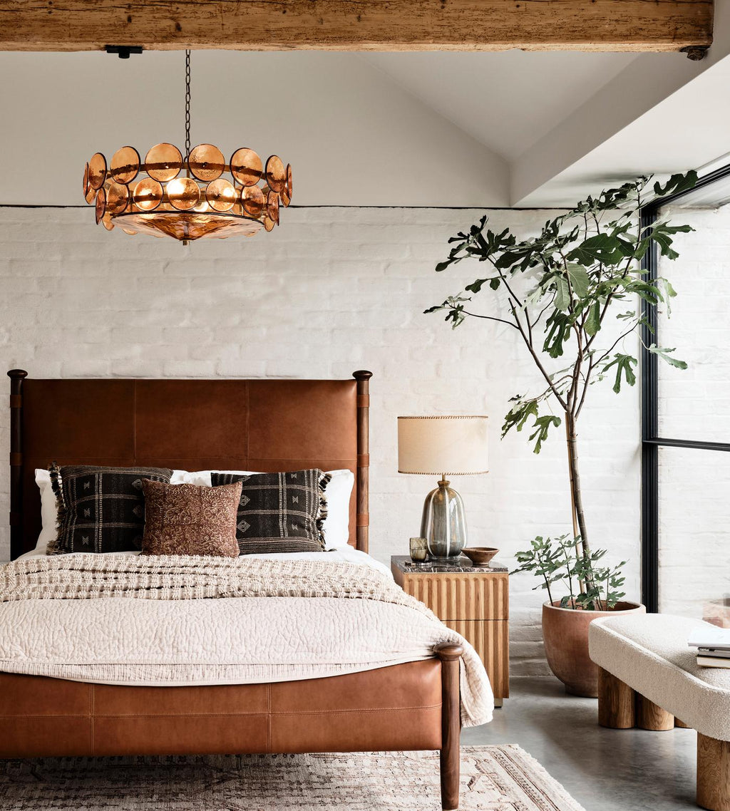 This Clérachic amber disc chandelier hangs in a loft bedroom with a white brick wall, a leather bed frame and a large potted plant.