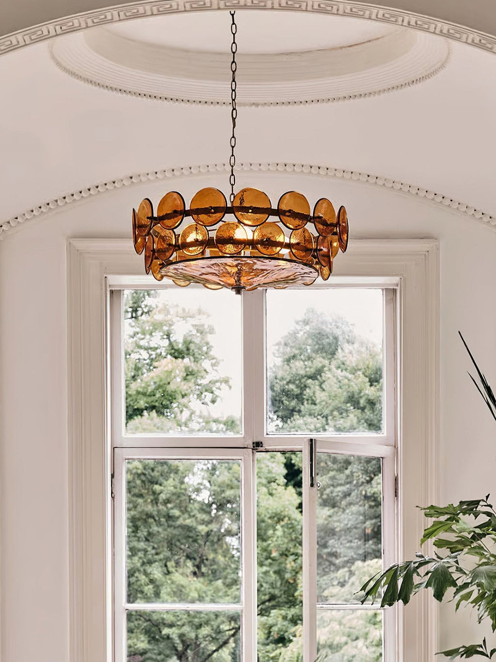Amber disc chandelier from Clérachic centred under an arched ceiling dome, in front of a tall window with green trees outside.