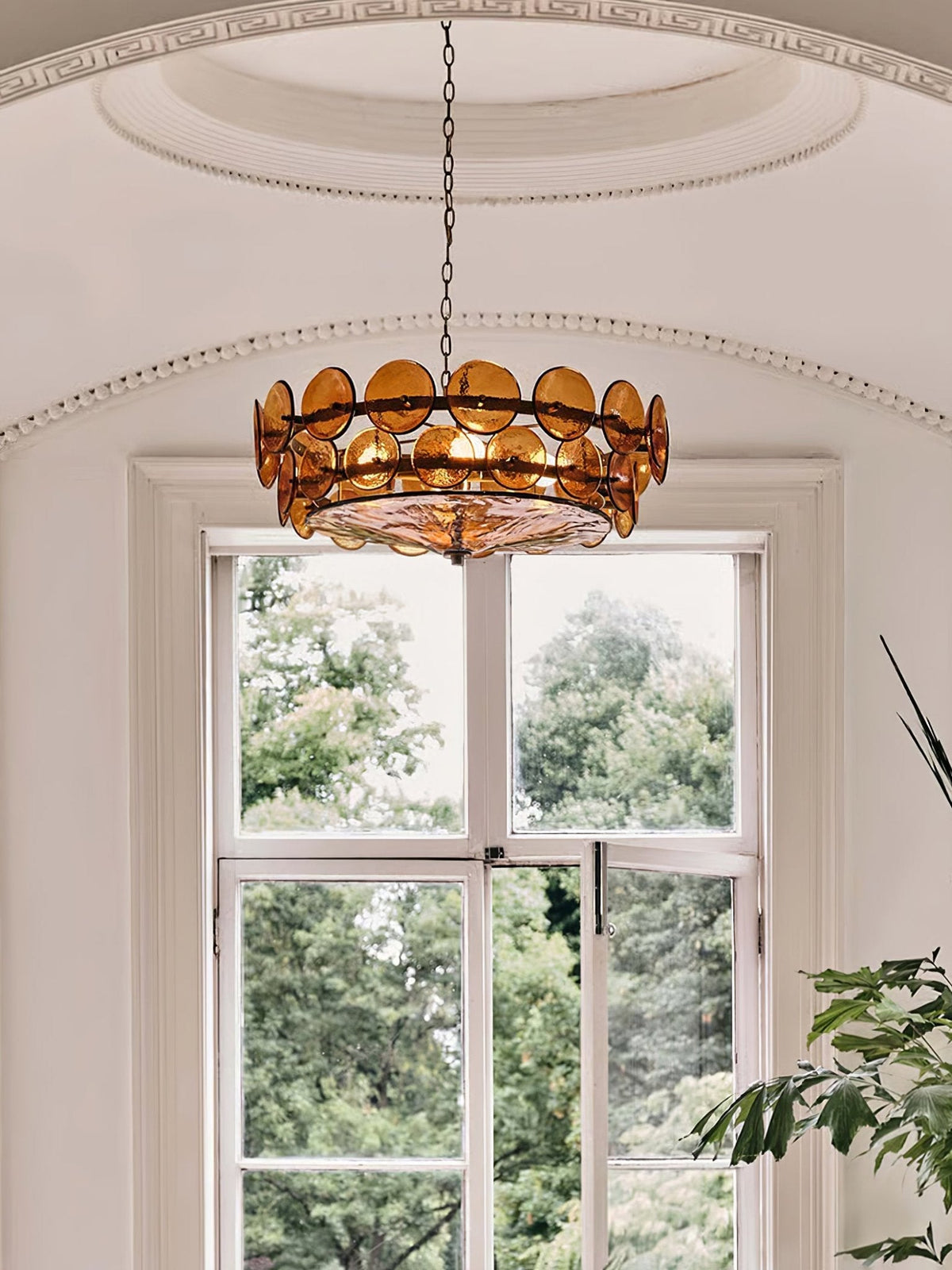 Amber disc chandelier from Clérachic centred under an arched ceiling dome, in front of a tall window with green trees outside.