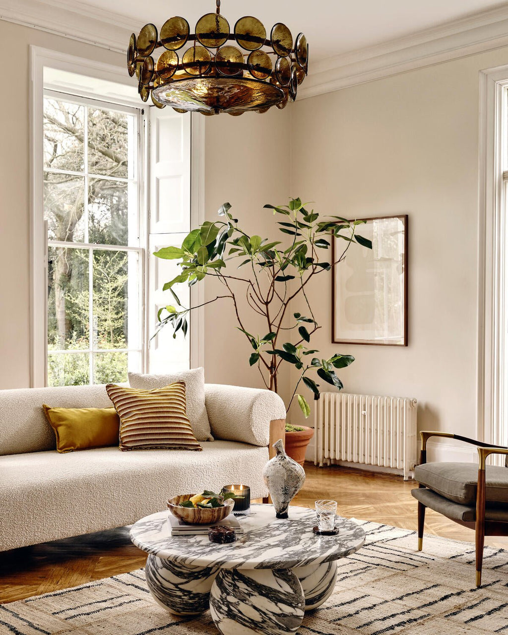 Green-gold disc chandelier from Clérachic high above a marble coffee table, a cream sofa and a leafy plant in a bright room.