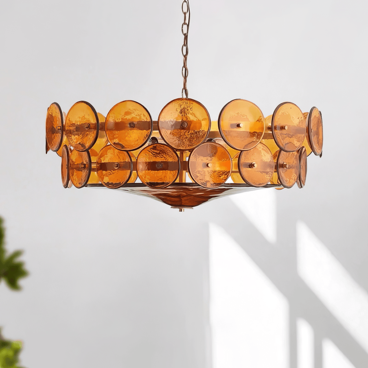 Amber glass disc chandelier from Clérachic hanging in front of a white wall with soft window shadows and a hint of leaves.