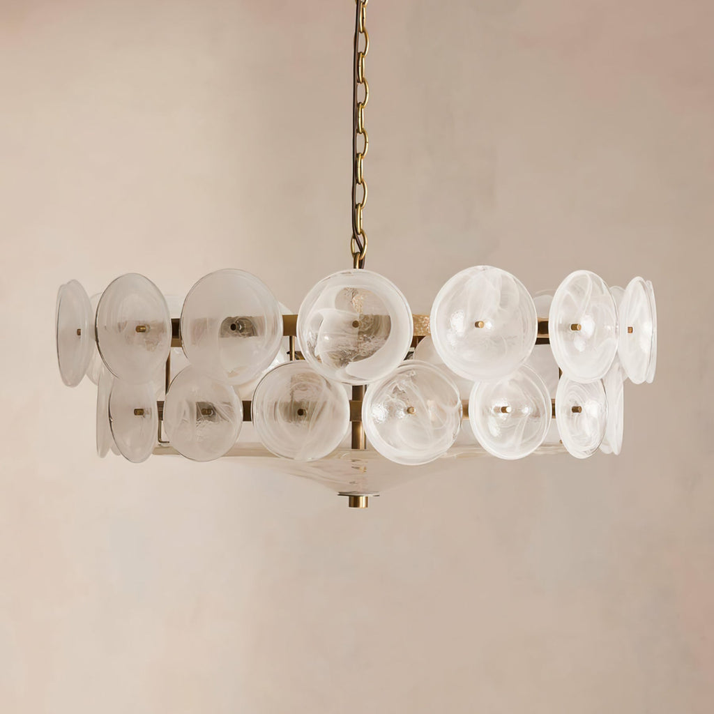 Frosted white swirled glass discs on a brass frame, this Clérachic chandelier hangs against a soft beige plaster wall.