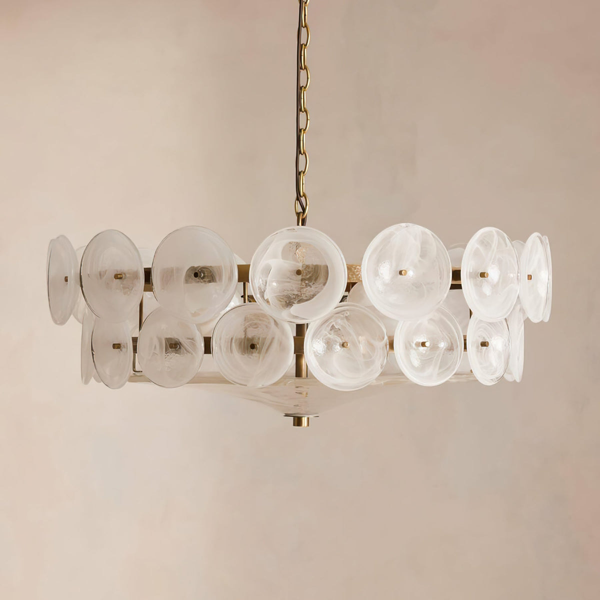 Frosted white swirled glass discs on a brass frame, this Clérachic chandelier hangs against a soft beige plaster wall.