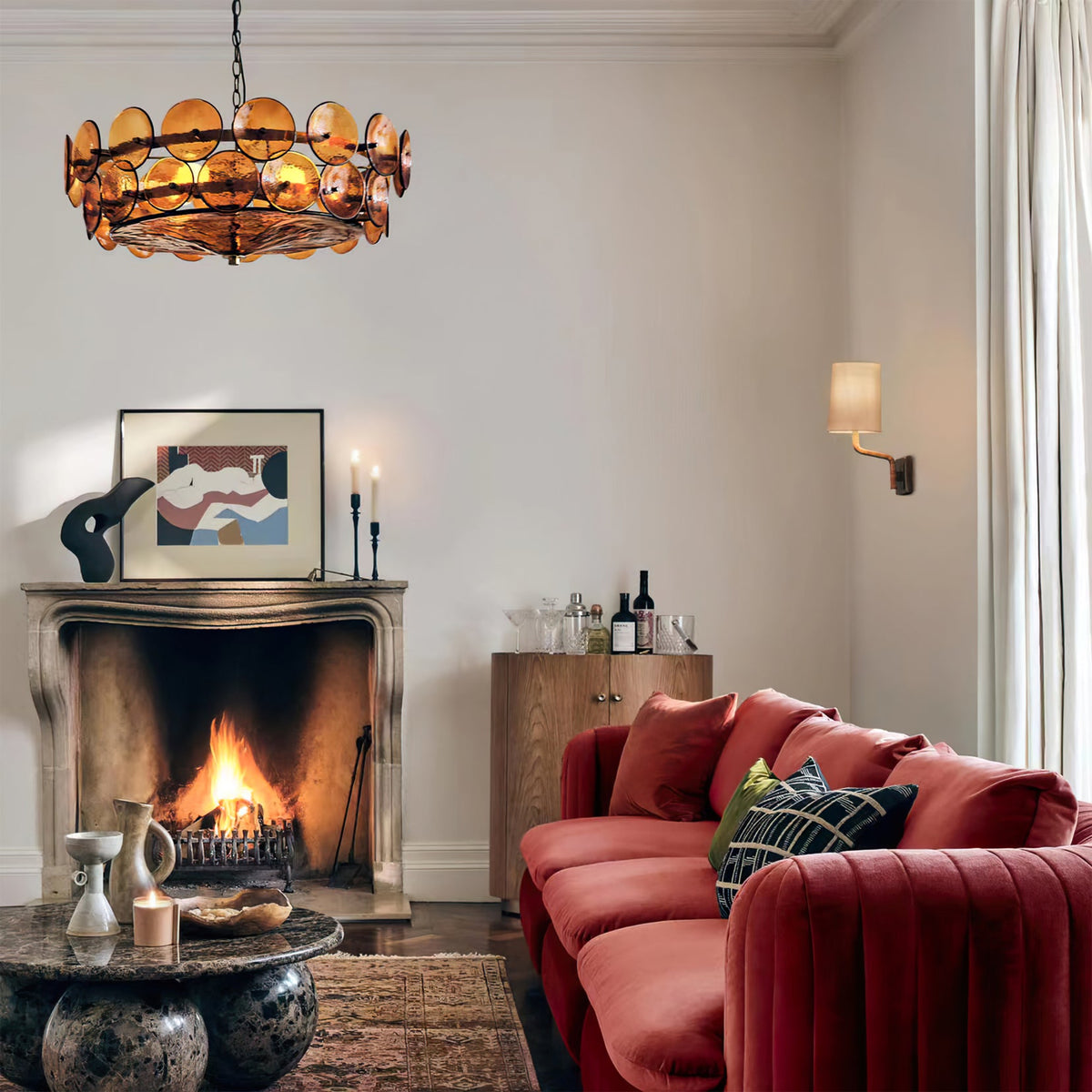 Amber disc chandelier from Clérachic above a fireplace and a red velvet sofa, with candles on the mantel and an abstract print.