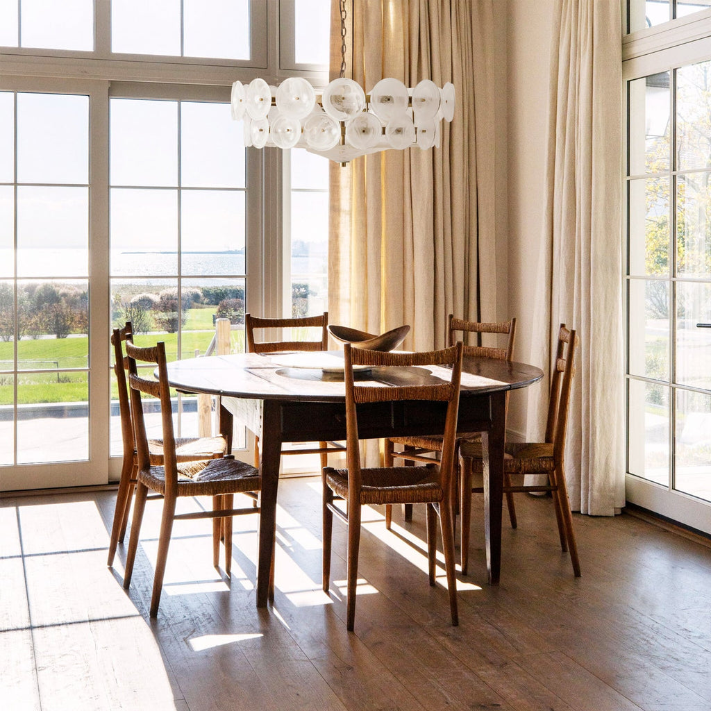 White glass disc chandelier from Clérachic above a dark oval dining table with rush-seat chairs, beside tall windows onto a lawn.