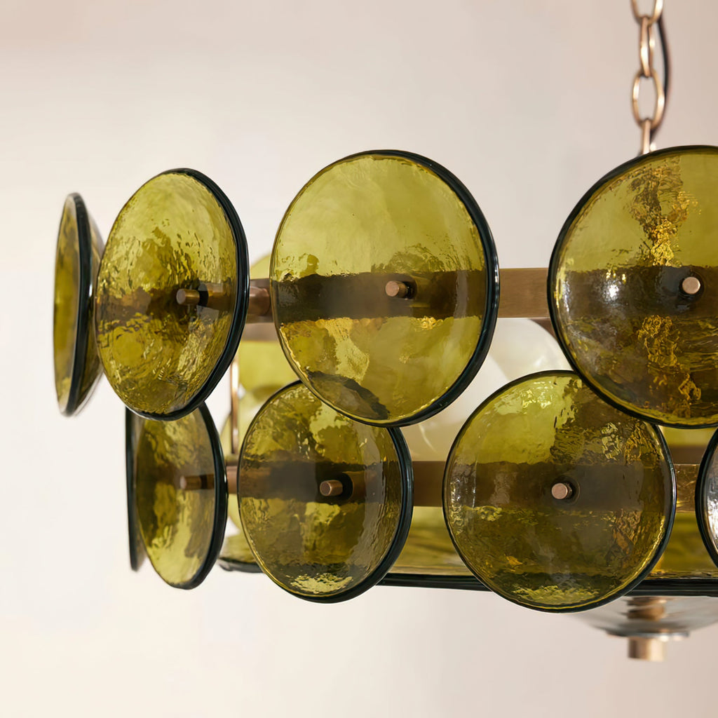 Detail of olive green glass discs with dark edges and brass studs, on a Clérachic chandelier, above a pale wall.