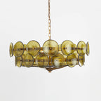 Green and gold Clérachic chandelier with olive discs and a long brass chain, hung against a pale grey studio wall.