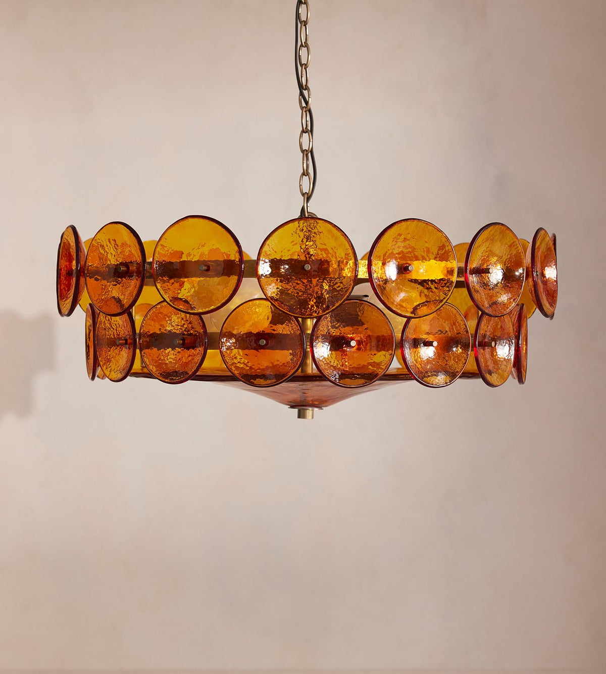 Lit amber disc chandelier from Clérachic against a soft pink-grey wall, with a dark chain and a ruby-toned bowl.