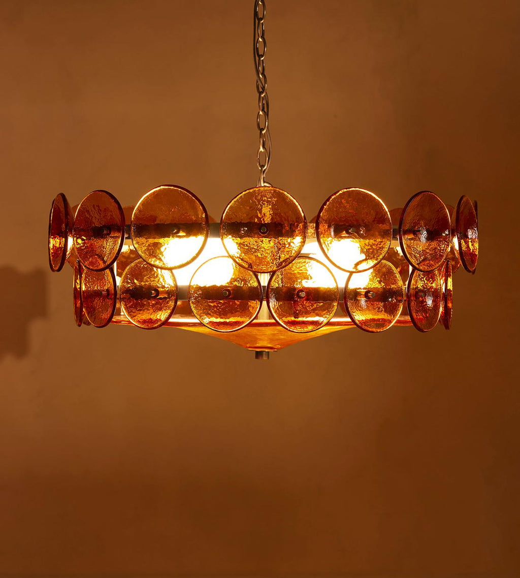 Amber glass disc chandelier from Clérachic lit bright, with the bulbs showing through the discs against a copper-brown wall.