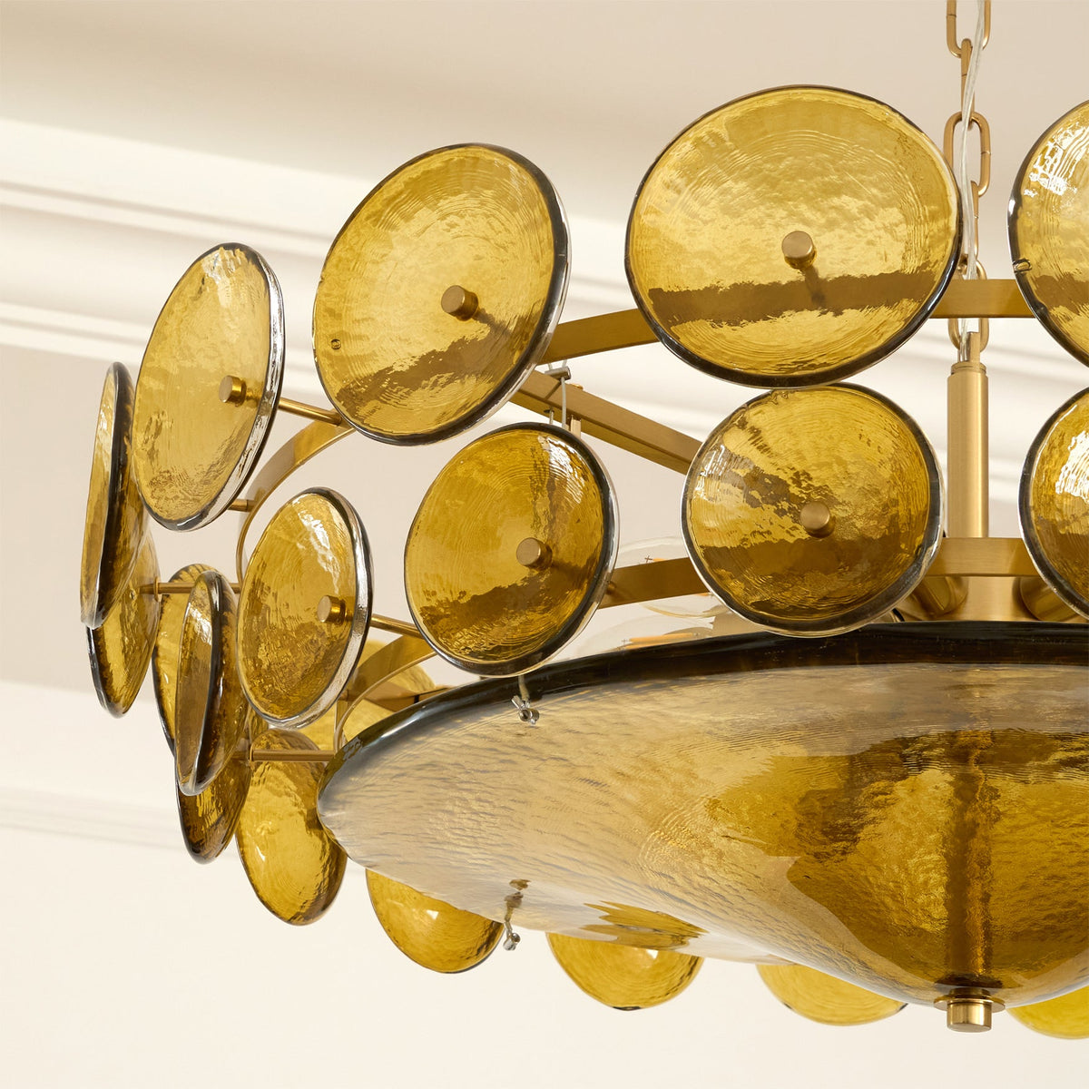 Underside of this Clérachic chandelier, with olive-gold discs on curved brass arms and a dark-rimmed glass bowl lit warm.
