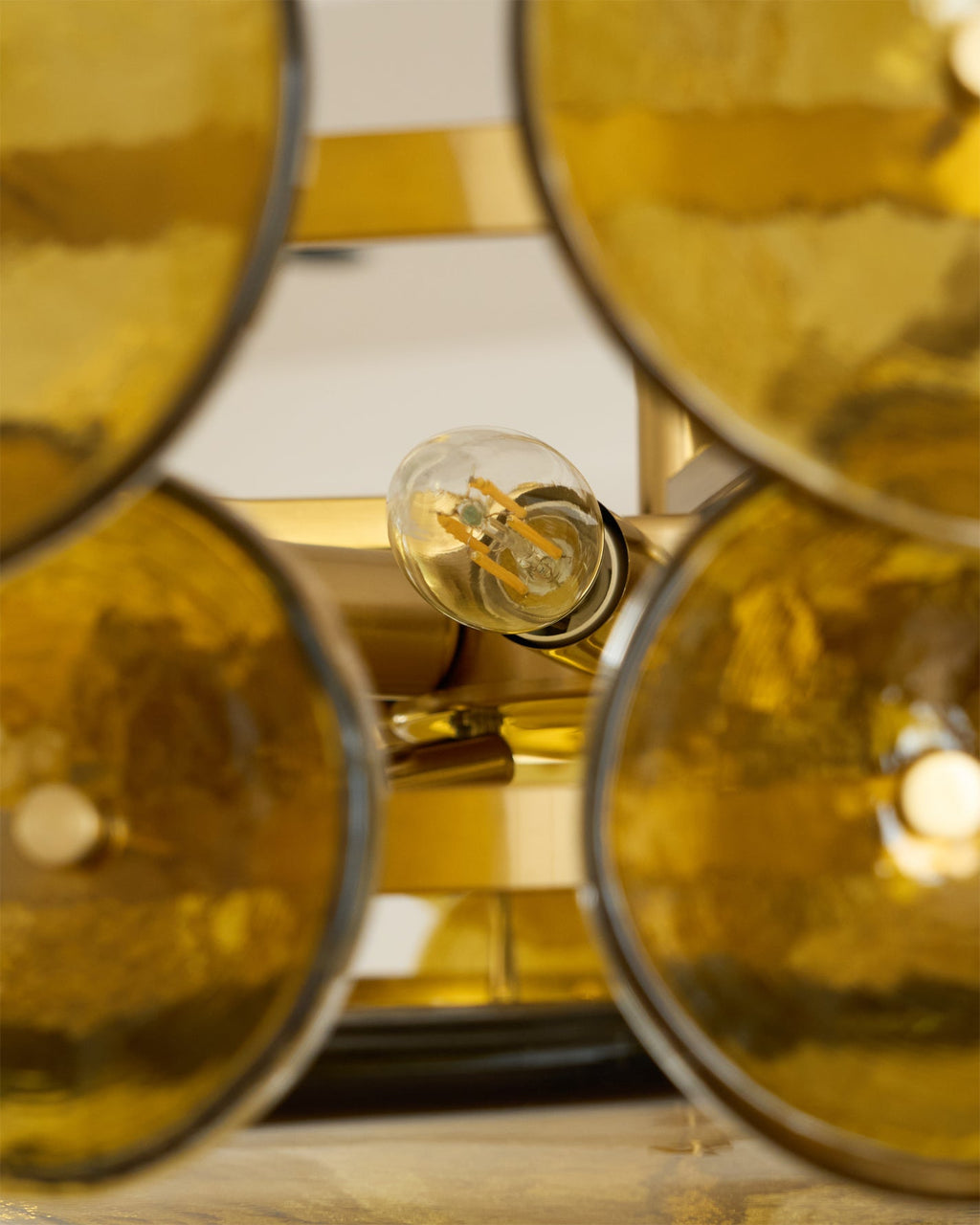 Tight crop of a filament bulb between olive-gold glass discs on the brass frame of this Clérachic chandelier.