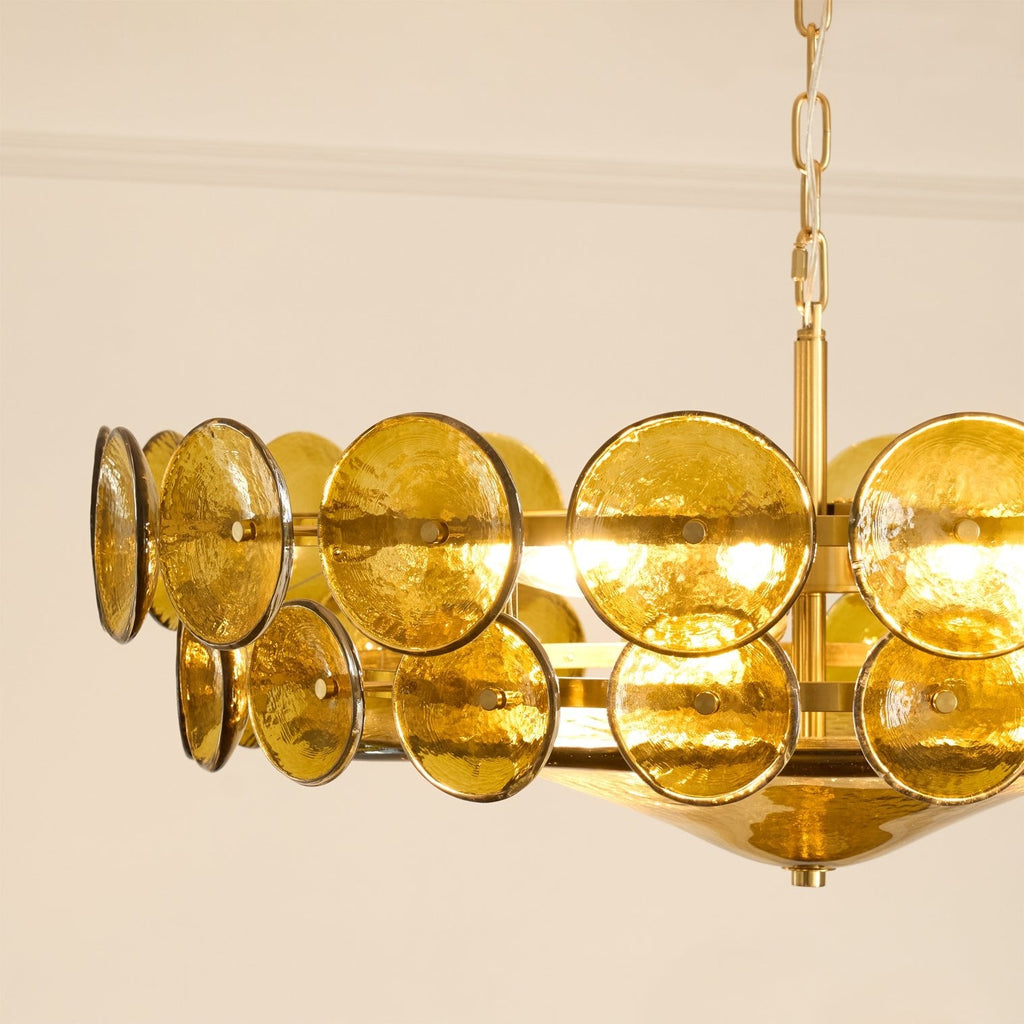 Close view of the green-gold glass discs and the brass rod and chain on a Clérachic chandelier, against a cream wall.