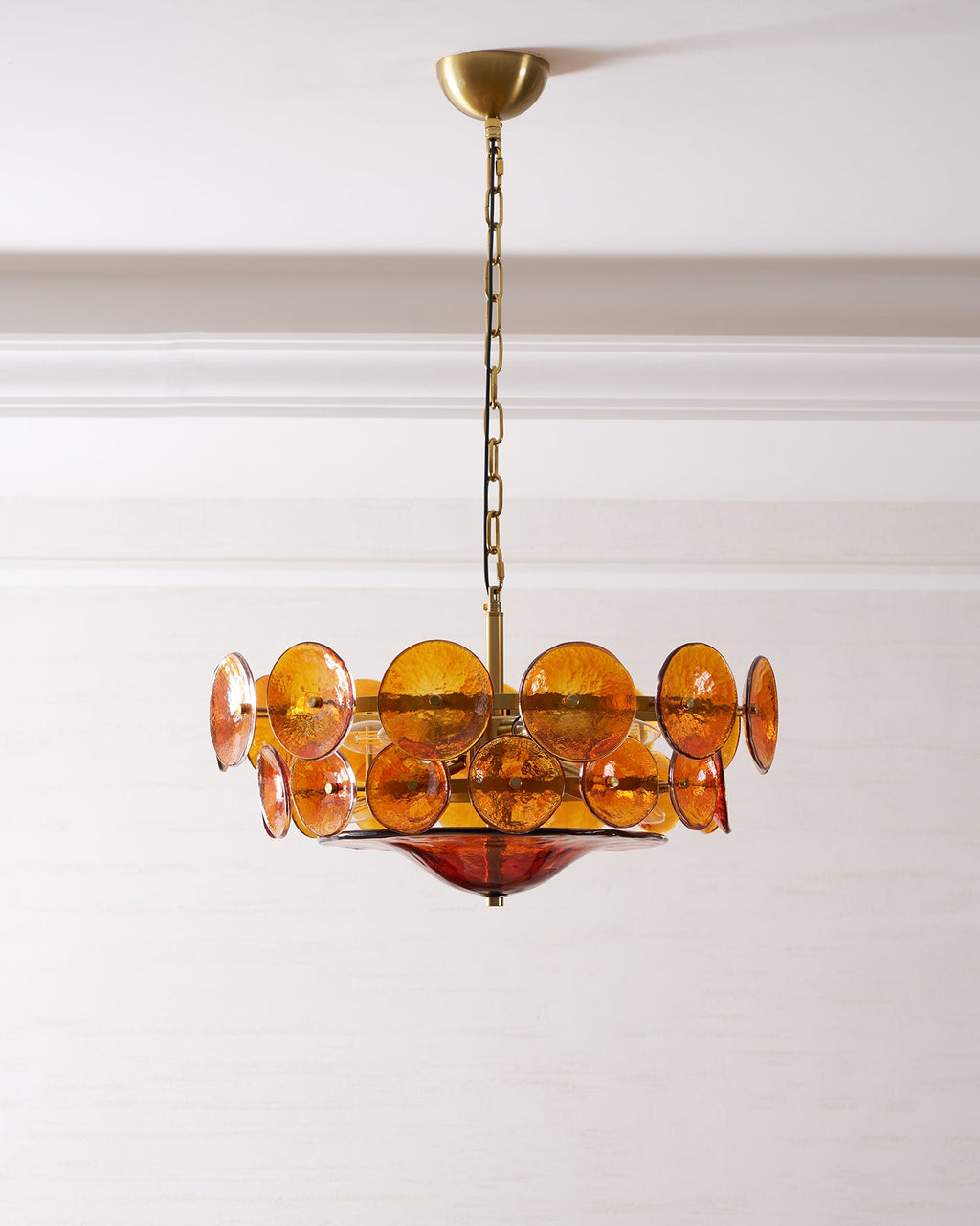 Medium amber glass disc chandelier from Clérachic on a gold canopy and chain, against a white panelled wall.