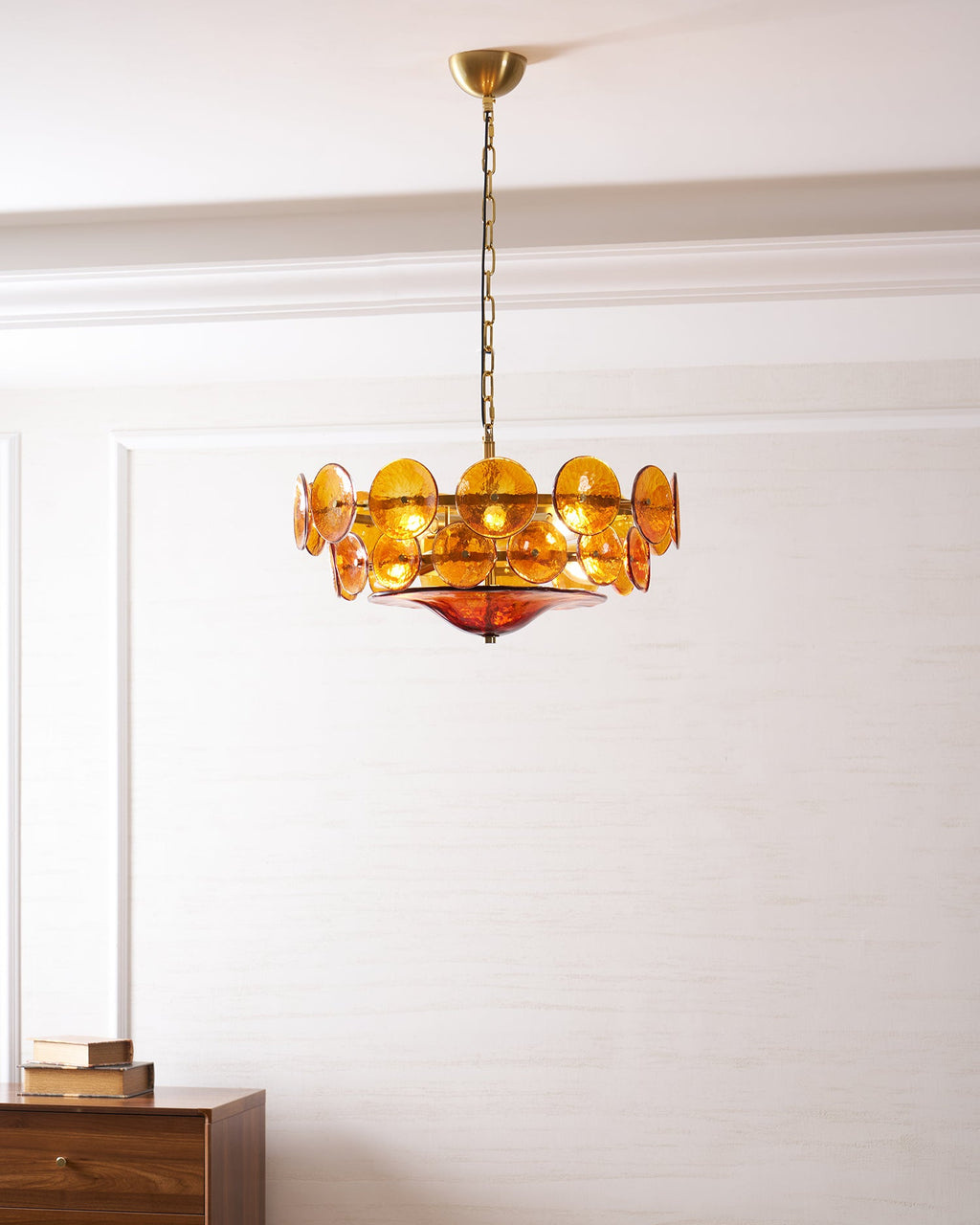Amber chandelier from Clérachic, lit, in a white room with a wooden sideboard and a stack of books at the left.