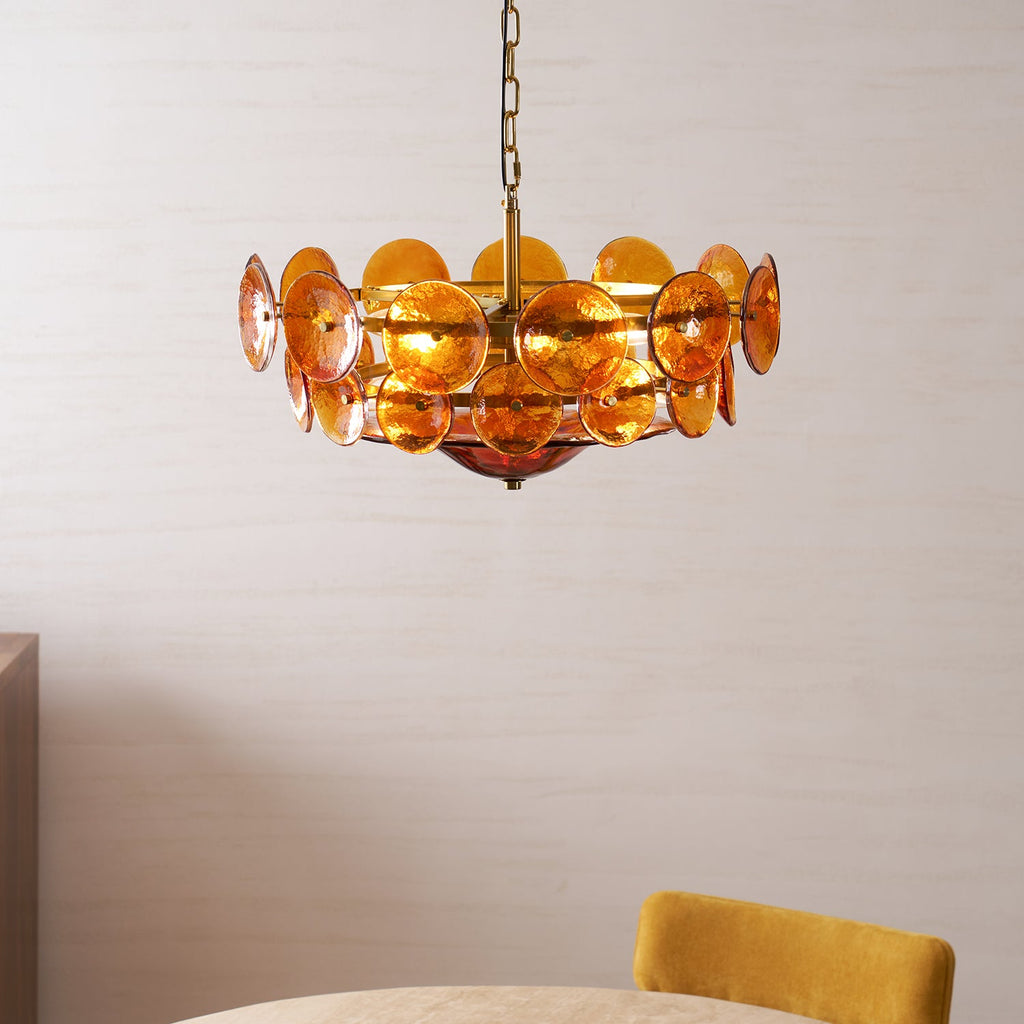 Lit amber glass disc chandelier from Clérachic above the edge of a dining table with a mustard-yellow chair, against a pale wall.