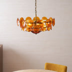 Lit amber glass disc chandelier from Clérachic above the edge of a dining table with a mustard-yellow chair, against a pale wall.