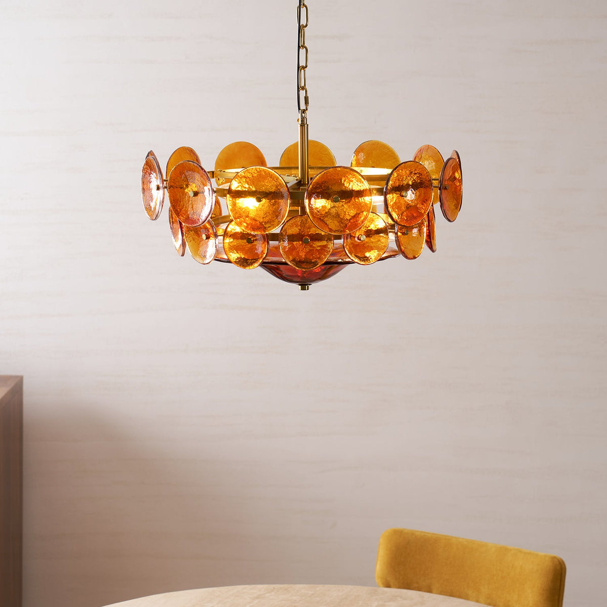 Lit amber glass disc chandelier from Clérachic above the edge of a dining table with a mustard-yellow chair, against a pale wall.