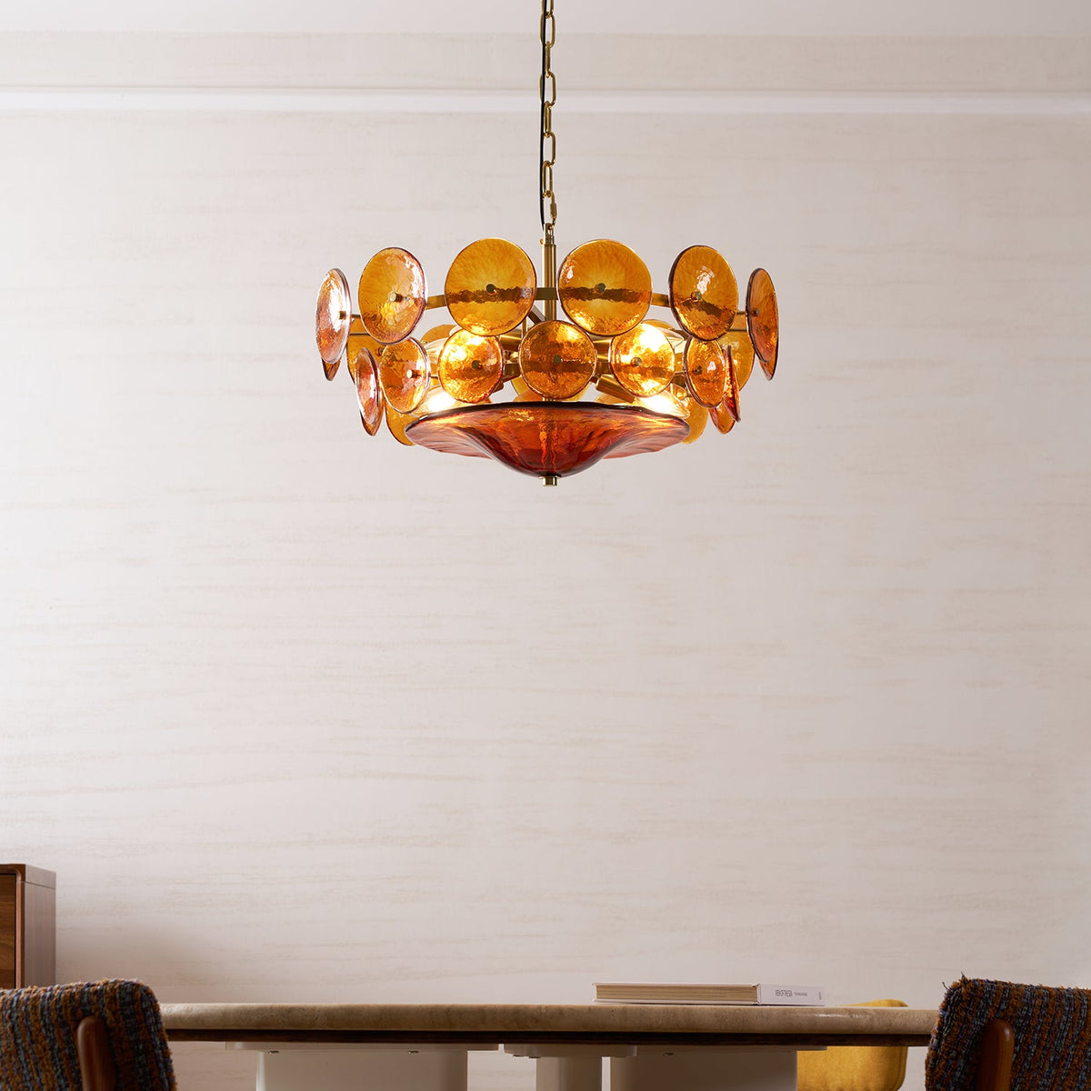 Amber disc chandelier from Clérachic over a round dining table with brown woven chairs, glowing against a pale textured wall.