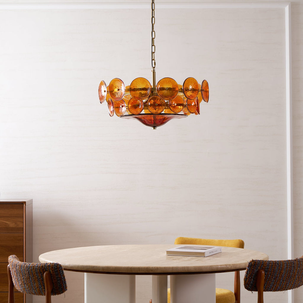 Lit amber disc chandelier from Clérachic over a round travertine table with a book, mustard and brown chairs, and a wooden cabinet.