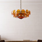 Larger view of the lit amber discs and ruby glass bowl on a Clérachic chandelier, hanging in a pale room above a low sofa.