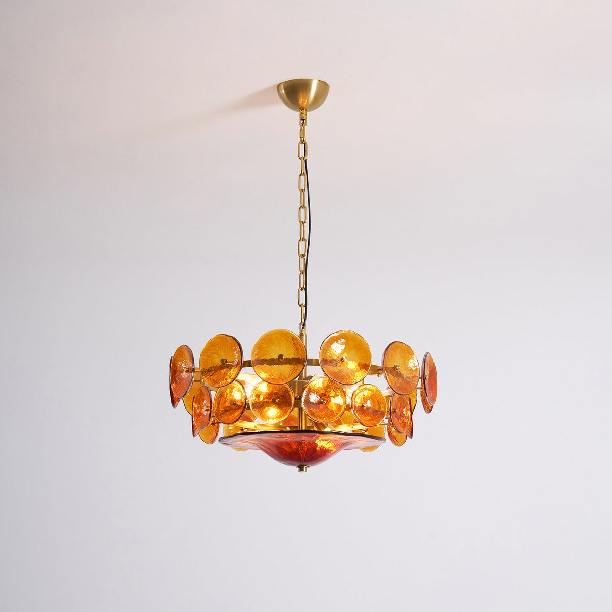 This Clérachic amber chandelier lit up with warm bulbs, hanging from a gold canopy in front of a pale grey wall.