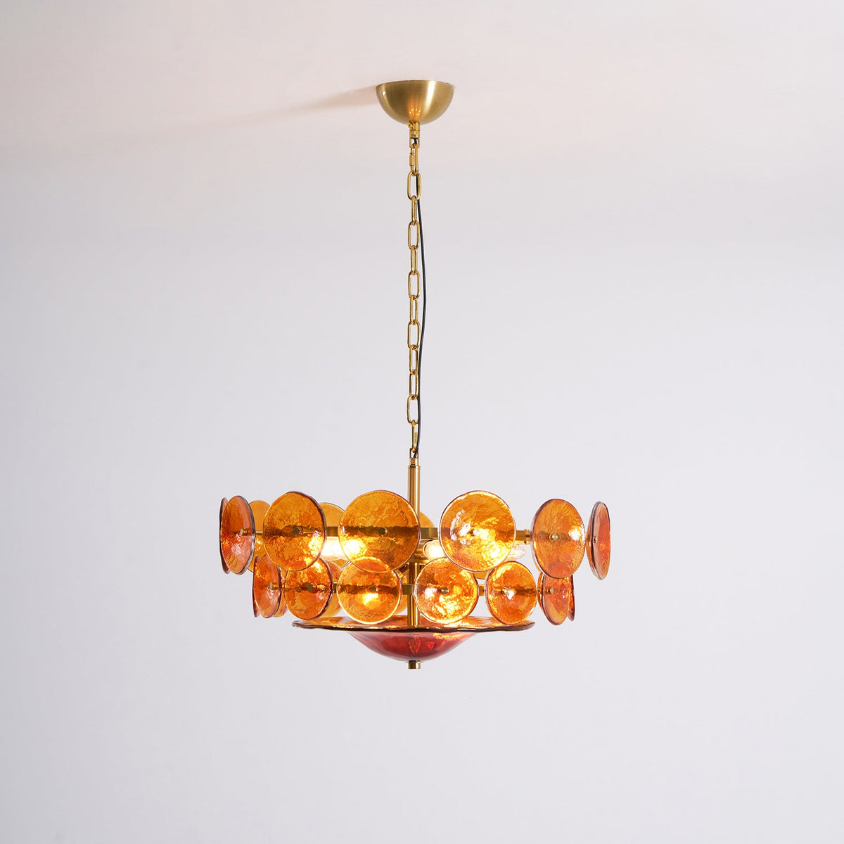 Close view of the lit amber disc chandelier from Clérachic, with its brass canopy and chain, against a white wall.