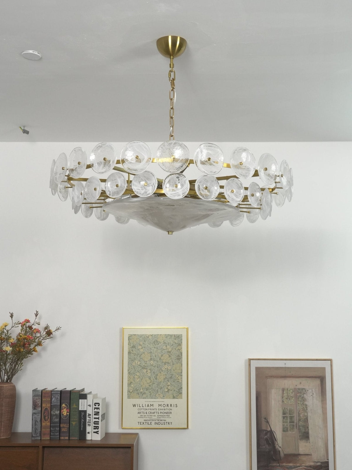 White glass disc chandelier from Clérachic hung in a plain white room, above a shelf with books and two framed prints.