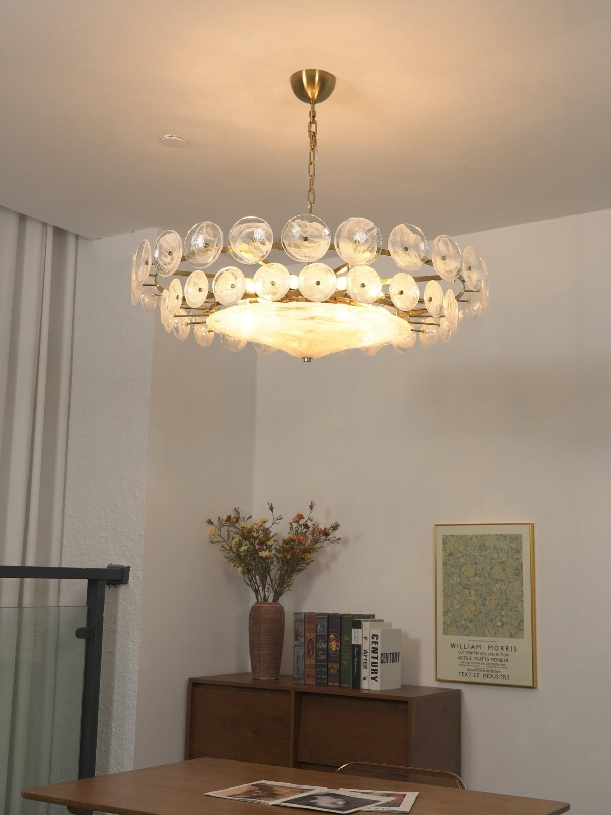 Lit white glass disc chandelier from Clérachic in a dining corner with a wooden table, a sideboard, books and a framed print.