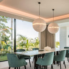 Two of this Clérachic pendant's spheres hang over a long white dining table with green velvet chairs, in front of tall glass doors to a garden.