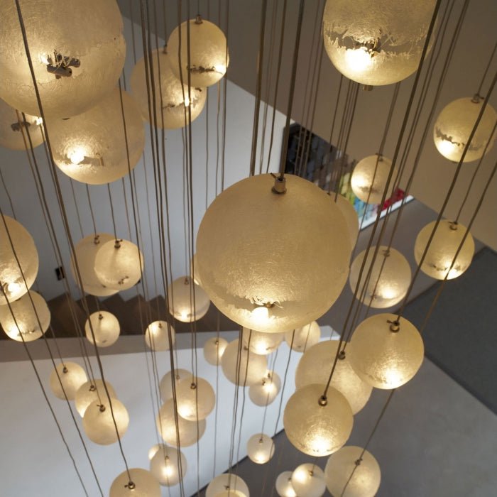 Looking down through a stairwell at many glowing spheres of different sizes on long cords, a cascade of this Clérachic pendant.
