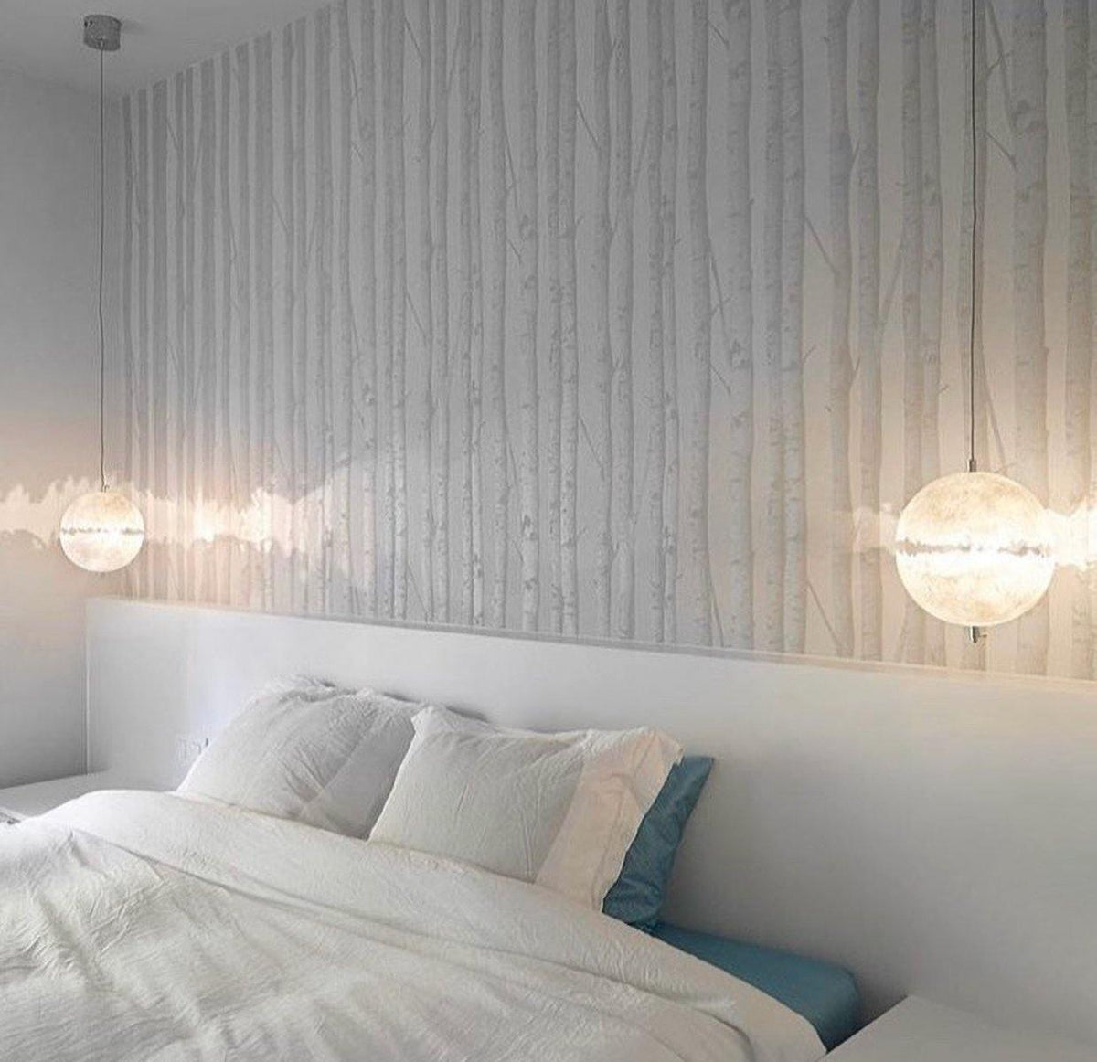 White sphere with a clear ragged band glowing above a bed, with a second sphere on the left, in a bedroom with a birch-pattern wall, shown on this Clérachic pendant.