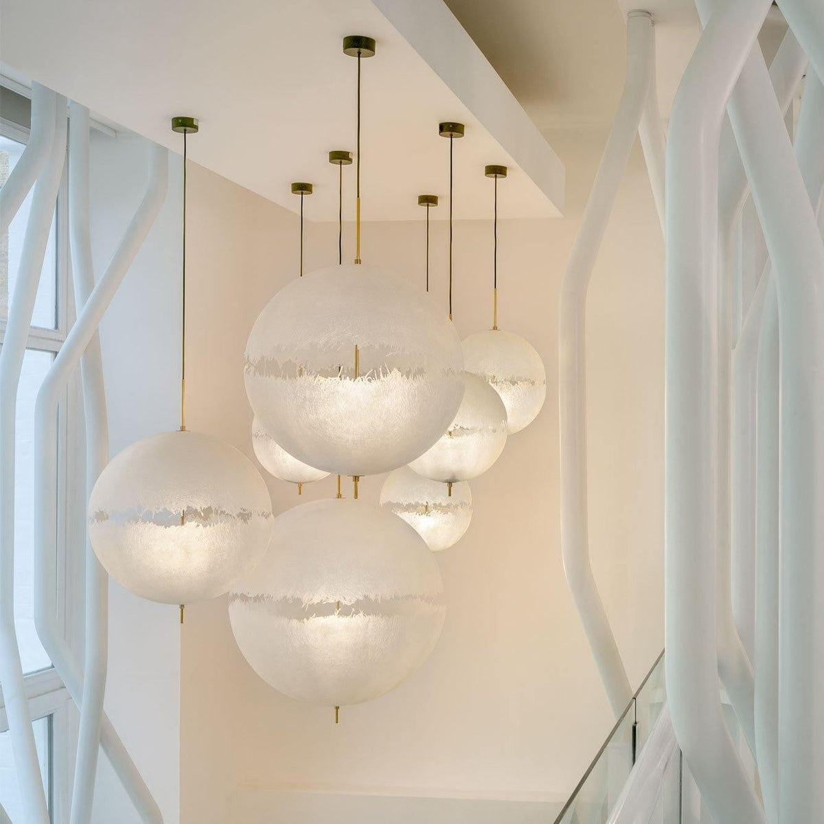 A cluster of this Clérachic pendant's spheres hangs from gold-toned stems in a bright stairwell beside white tree-like supports.