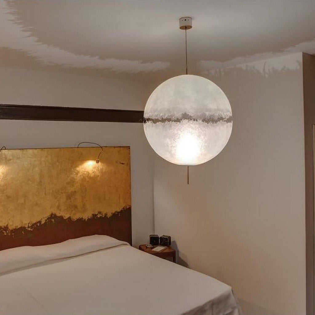 One lit sphere with a ragged band hangs above a bed with a gold-leaf headboard, from this Clérachic pendant on a pale cord.