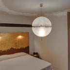 One lit sphere with a ragged band hangs above a bed with a gold-leaf headboard, from this Clérachic pendant on a pale cord.