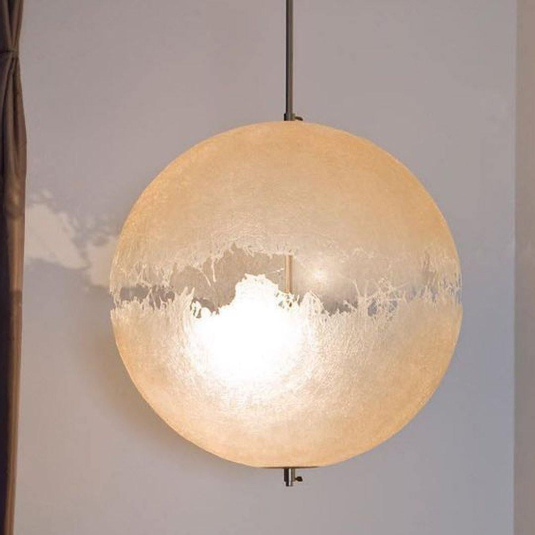Large lit sphere of this Clérachic pendant, glowing amber, with its torn-edge clear band and a small metal tip at the base.