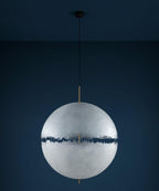 Unlit pale sphere with a dark jagged band on a thin black cord, this Clérachic pendant against a deep navy wall.