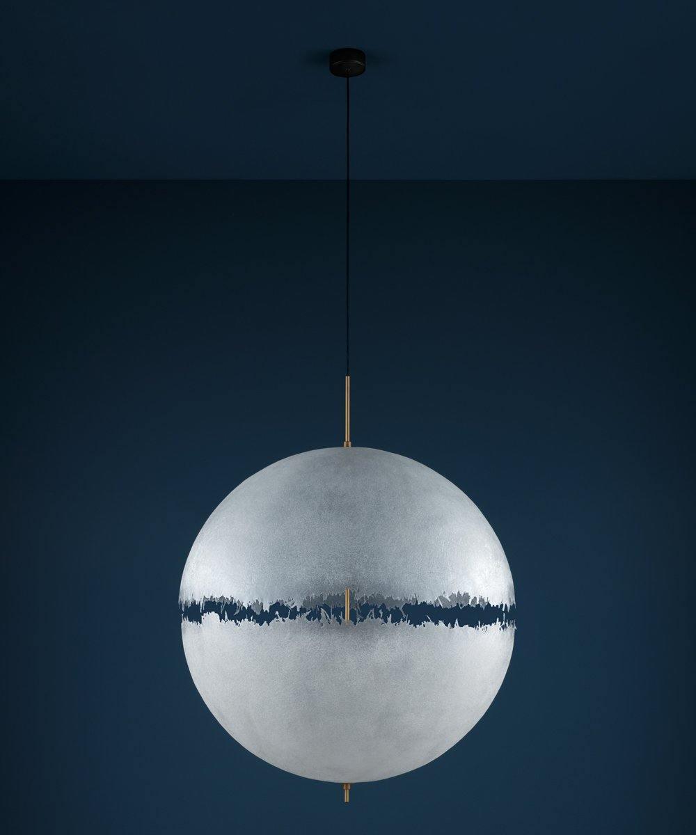 Unlit pale sphere with a dark jagged band on a thin black cord, this Clérachic pendant against a deep navy wall.