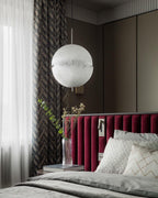 This Clérachic pendant hangs by a curtain and a bed with a deep red headboard in a calm, dark-toned bedroom.