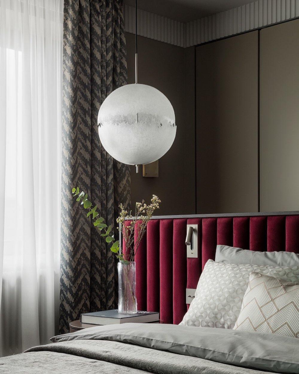 This Clérachic pendant hangs by a curtain and a bed with a deep red headboard in a calm, dark-toned bedroom.