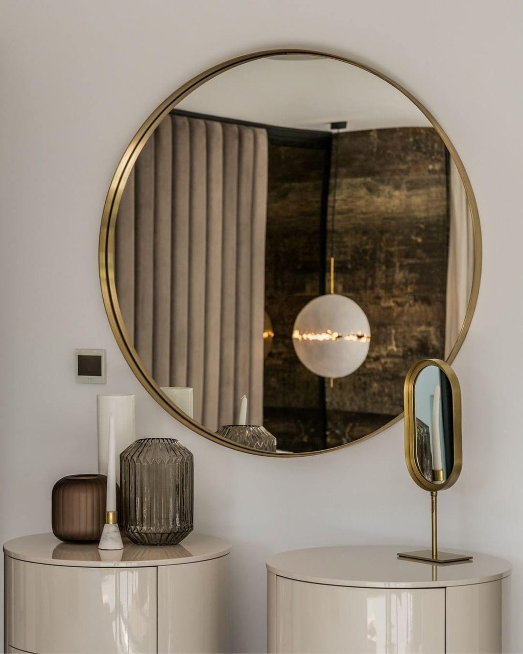 A single small sphere of this Clérachic pendant reflected in a round brass-framed mirror above two cream bedside drawers.