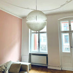 A large white sphere from this Clérachic pendant hangs in a high room with a pink wall and tall windows and a stone-toned sofa.