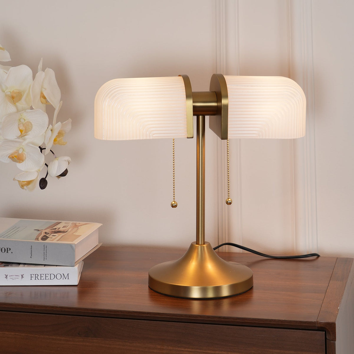 This Clérachic gold table lamp with white ribbed shades sits on a walnut chest, beside white orchids and a stack of books.