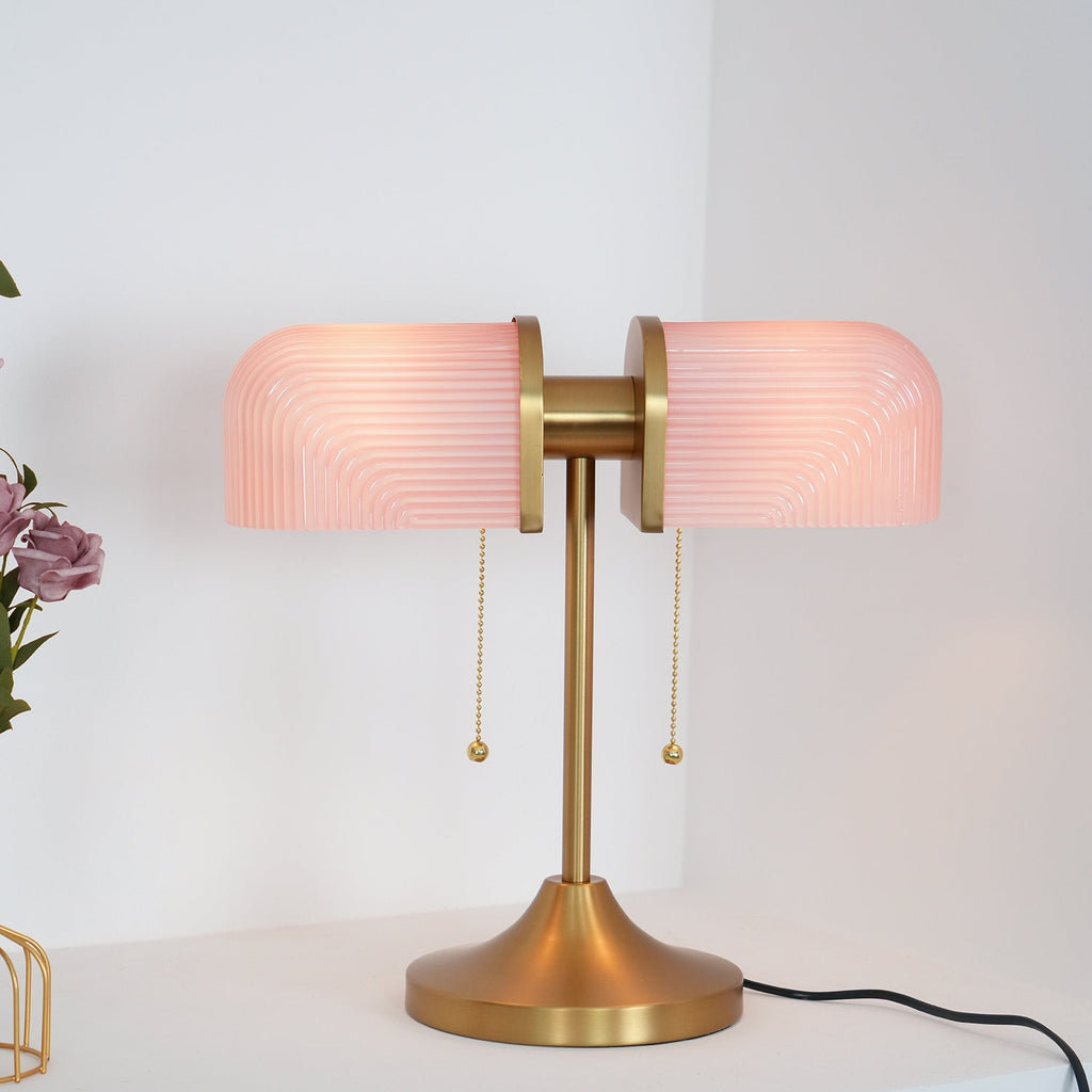 Shown here, a Clérachic gold table lamp with pink ribbed glass shades glowing, on a white shelf beside a wire vase and its black cord.