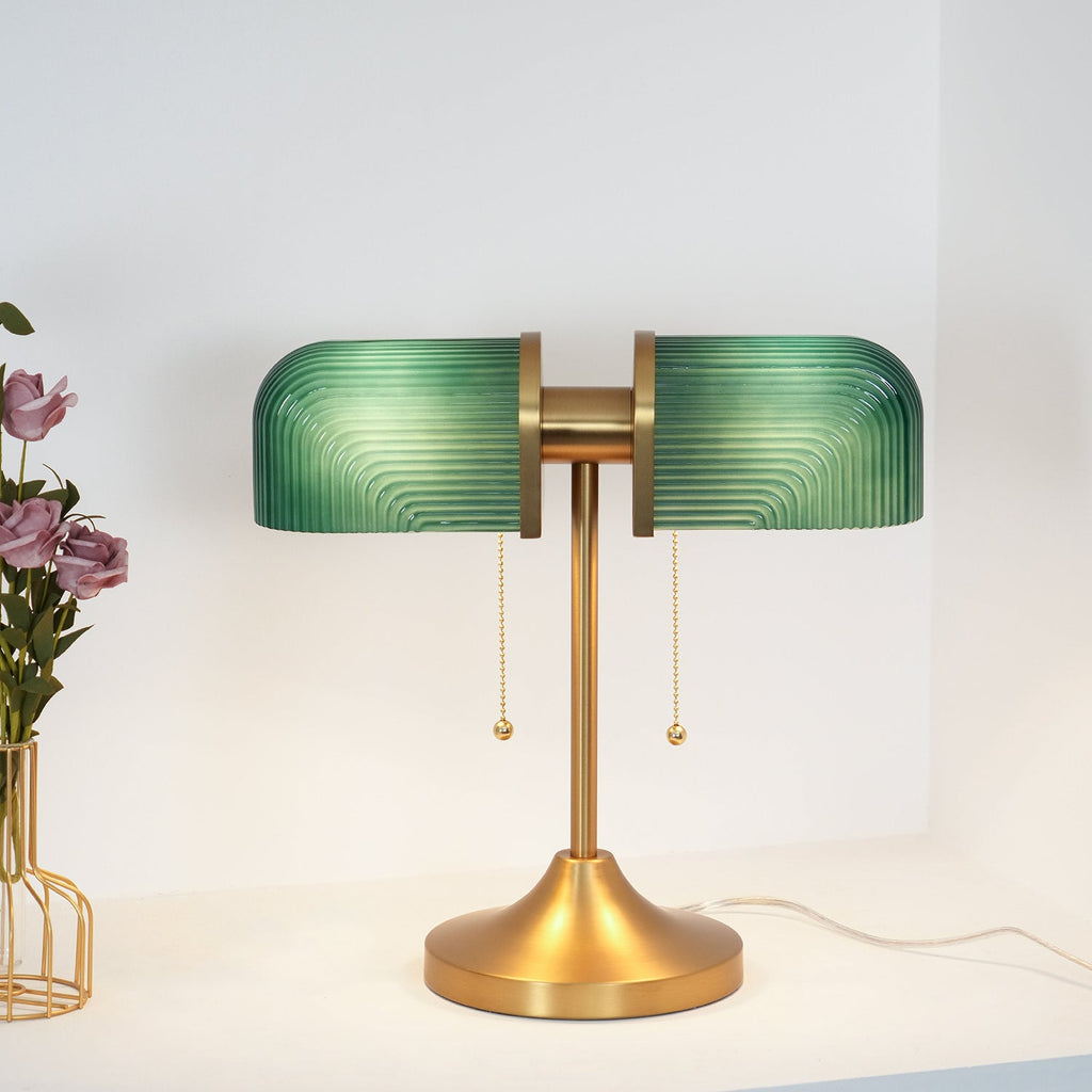 This Clérachic gold table lamp has deep green ribbed glass shades, with two brass pull chains, on a white shelf by pink roses.
