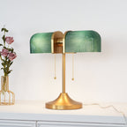 A green-shaded Clérachic gold table lamp stands on a white cabinet, a vase of dusky pink flowers at its left.