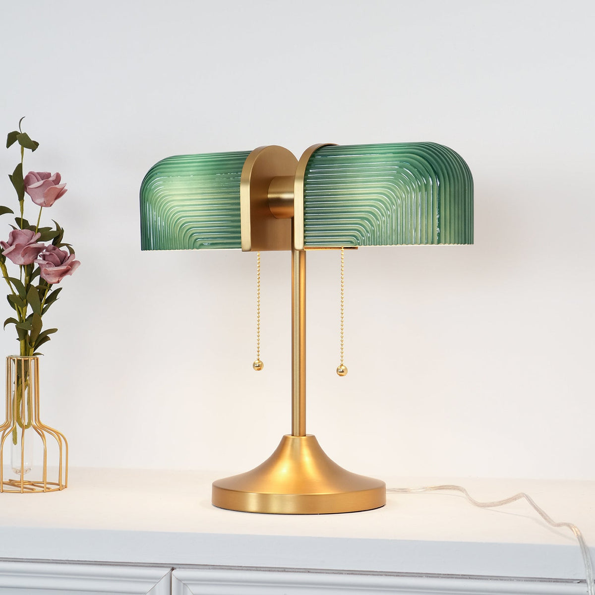 A green-shaded Clérachic gold table lamp stands on a white cabinet, a vase of dusky pink flowers at its left.
