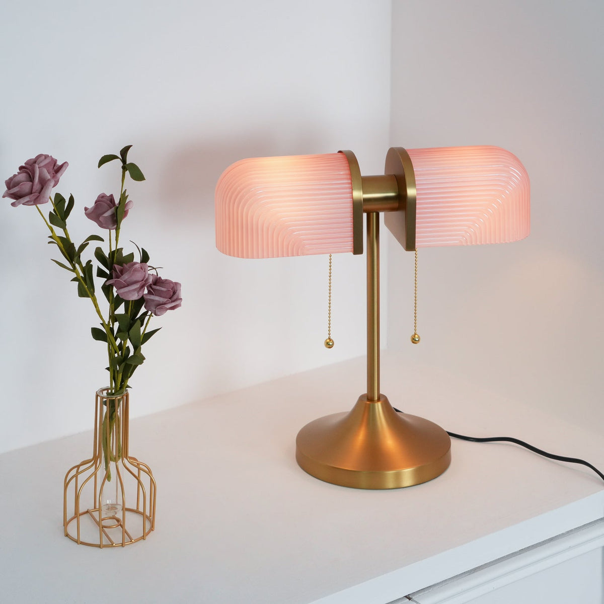 This Clérachic gold table lamp with pink ribbed shades glows beside a wire vase of mauve roses on a white cabinet.
