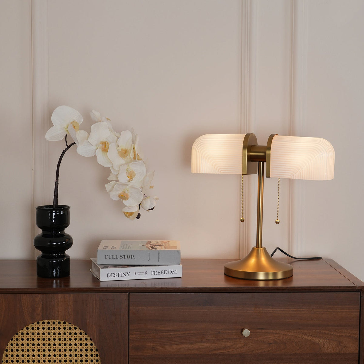 In this photo, a Clérachic gold table lamp with white ribbed shades lit on a walnut chest of drawers, beside a black glass vase and white orchids.